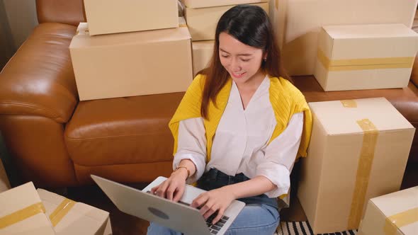 Young Asian woman sit on floor and lean to sofa with many boxes or carton and use laptop