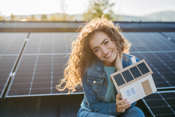 Portrait of young woman on roof with solar panels, holding model of house with photovoltaics. - Stock Photo - Images