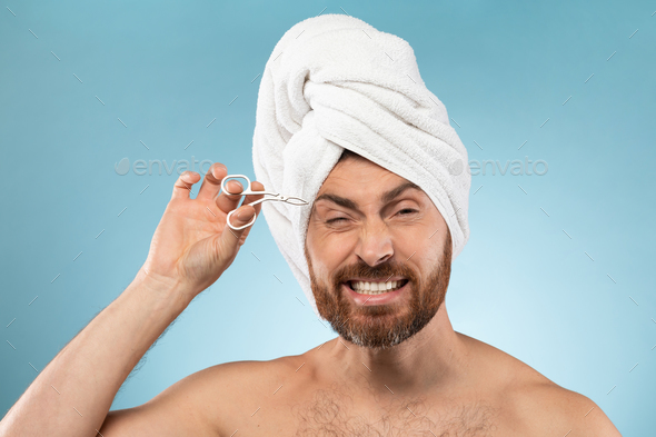 Funny emotional bearded man plucking his eyebrows and using tweezers, standing with towel on head