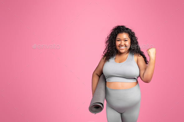 Excited black oversize lady in sportswear making win sign, celebrating success, holding mat, pink