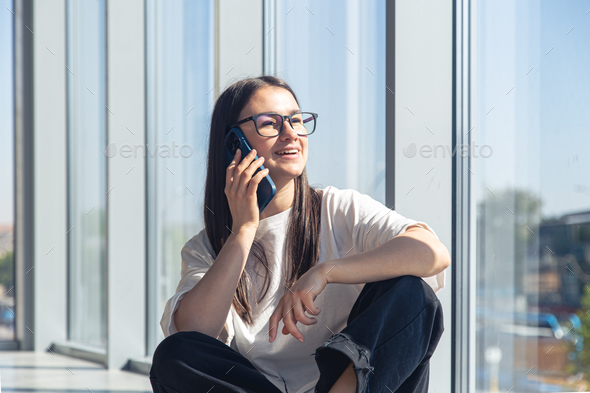 A young woman in glasses is talking on a smartphone near the window. - Stock Photo - Images