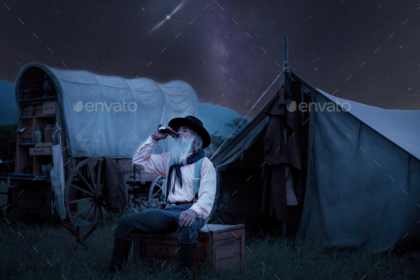 Oldest smart cowboy wearing western style with cowboy hat hold a cup drinking coffee on fire camp ea