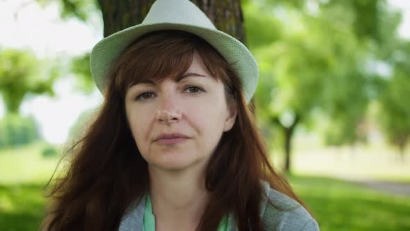 Portrait of a Woman in a Hat Near the Tree in the Park Looking at the Camera