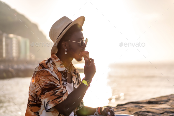 Sunset of a black ethnic man enjoy summer vacation on the beach eating an ice cream with a hat - Stock Photo - Images