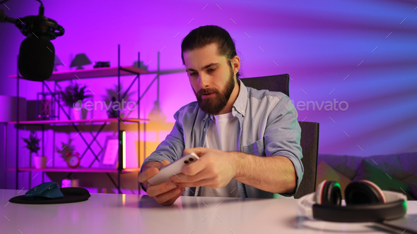 Gamer in neon room. A bearded guy sits at a table and plays a video game on his smartphone. Mobile