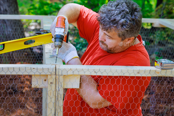 Worker uses screwdriver to wooden domestic chicken coop on farm that contains metal grid for - Stock Photo - Images