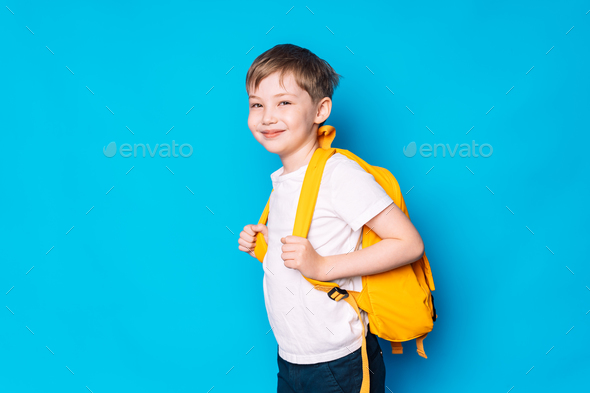 Schoolboy with yellow backpack stands sideways against blue background - Stock Photo - Images