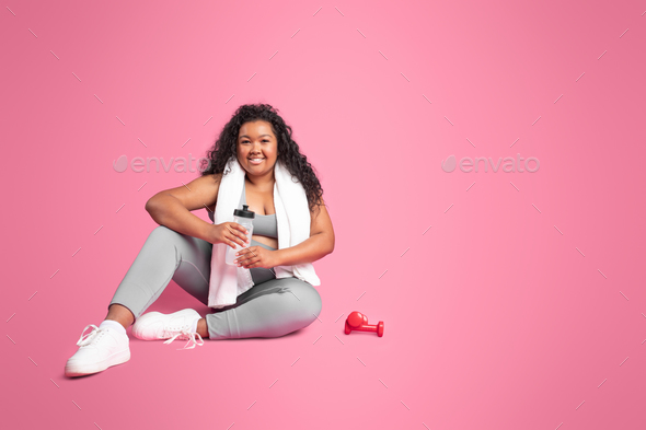 Happy black body positive lady in sportswear with dumbbells and bottle of water resting on pink