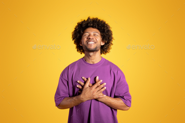 Happy satisfied mature black curly man in purple t-shirt with closed eyes presses hands to chest
