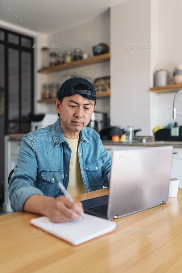 Asian man taking notes while working from the kitchen - Stock Photo - Images
