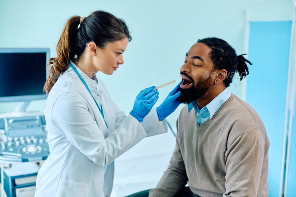 Female doctor using tongue depressor while checking black man\'s throat at medical clinic.