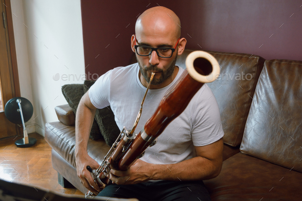front view of young classical music composer man, playing bassoon reading sheet music at home