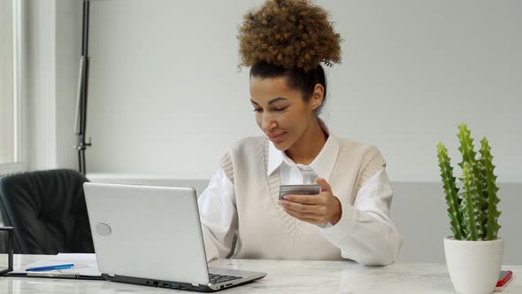AfricanAmerican Woman Holding a Credit Card Sitting at a Table with a Laptop Shopping Online Making alt