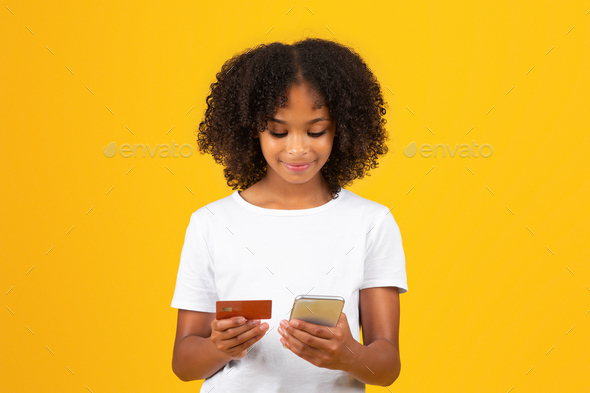Happy adolescent curly black girl in white t-shirt uses credit card and smartphone for online - Stock Photo - Images