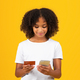 Happy adolescent curly black girl in white t-shirt uses credit card and smartphone for online - PhotoDune Item for Sale