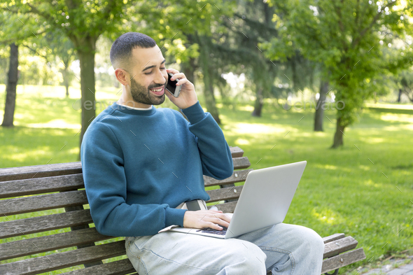 Young Caucasian man multitasking outdoors with laptop and phone - Stock Photo - Images