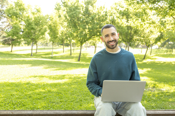 Young Caucasian happy and smiling man working on laptop outdoors, looking at the camera - Stock Photo - Images