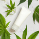 Blank Cream tube near green cannabis leaves on white table, top view. Cosmetic Mockup - PhotoDune Item for Sale
