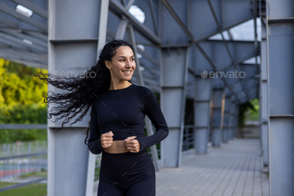 Young beautiful hispanic woman with curly hair jogging near stadium, woman in tracksuit smiling - Stock Photo - Images