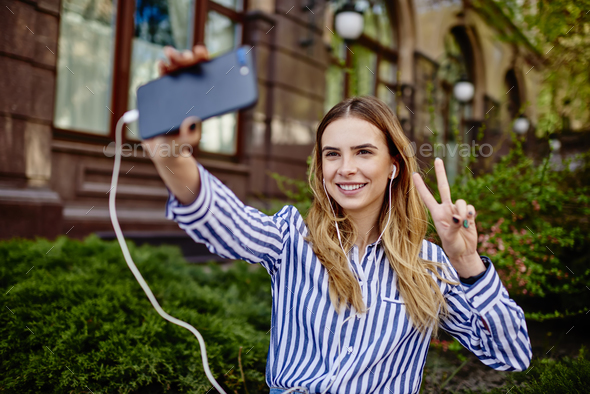 Cheerful young lady showing victory sign while making selfie outdoors