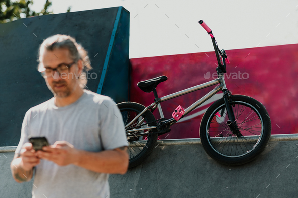 A bmx bike in a skatepark with a mature urban tattooed man using his phone in a blurry foreground. - Stock Photo - Images