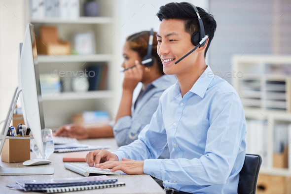 Happy young asian male call centre telemarketing agent talking on a headset while working on a comp - Stock Photo - Images