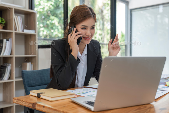 Cheerful young beautiful business woman talking on mobile phone and using laptop with smile while - Stock Photo - Images