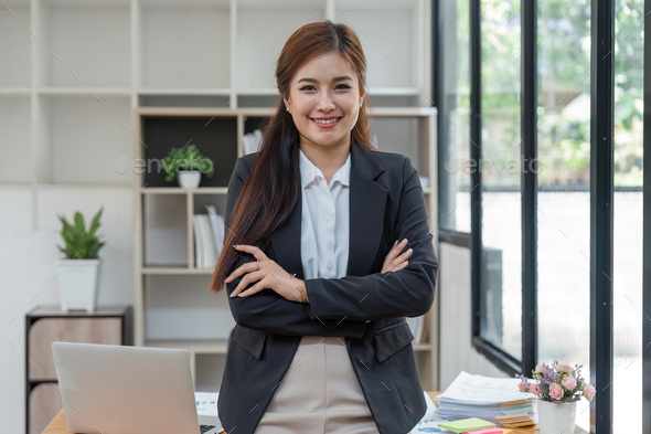 Young successful woman entrepreneur or an office worker stands with crossed arms near a desk in a - Stock Photo - Images