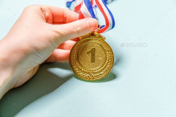 Closeup of a hand holding the first place gold trophy medal isolated on pale greenish background.