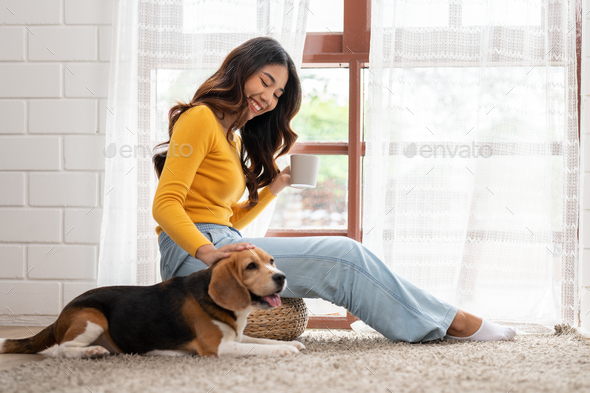 Asian young woman with her beagle dog relaxing and drinking coffee sitting in living room at home