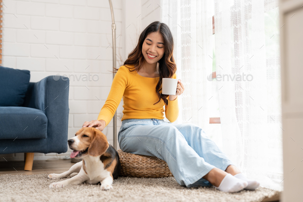 Asian young woman with her beagle dog relaxing and drinking coffee sitting in living room at home