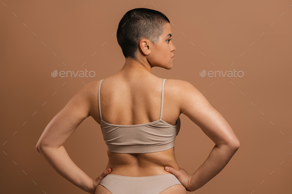 Rear view of shaved head woman in underwear holding hands on hip against grey background