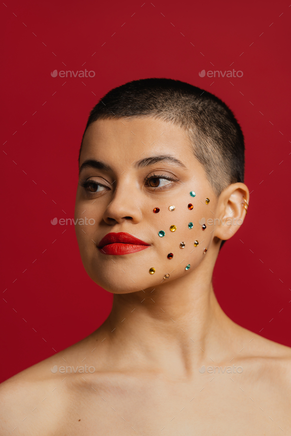 Beautiful young shaved head woman with shiny crystals over her face standing against red background