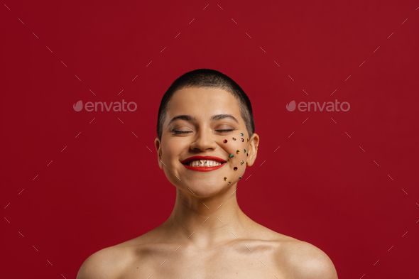 Happy young shaved head woman with shiny crystals over her face standing against red background