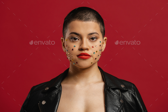 Beautiful shaved head woman with shiny crystals over her face standing against red background