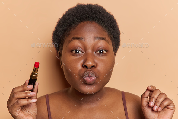 Beauty and cosmetics concept. Plump dark skinned female model keeps lips folded holds red lipstick
