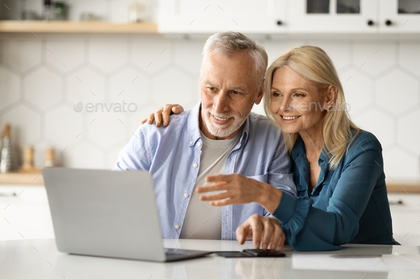 Happy Senior Couple Paying Taxes Online With Laptop In Kitchen