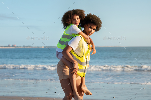 Happy young curly black lady volunteer in vest holds small child on her back, cleaning up garbage on - Stock Photo - Images