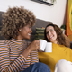 Multiethnic couple taking a coffee break sitting in the floor of a living room. Two cheerful women - PhotoDune Item for Sale