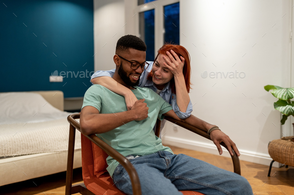 Smiling black man enjoy on armchair communication with hugging european laughing girlfriend at home - Stock Photo - Images