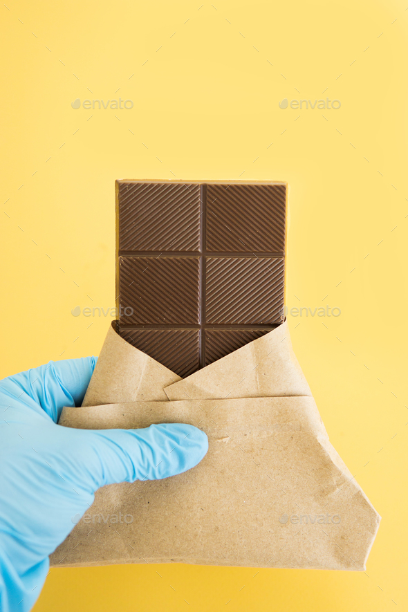 Bar of milk chocolate in a wrapper is held by a hand in a medical glove. World chocolate day.