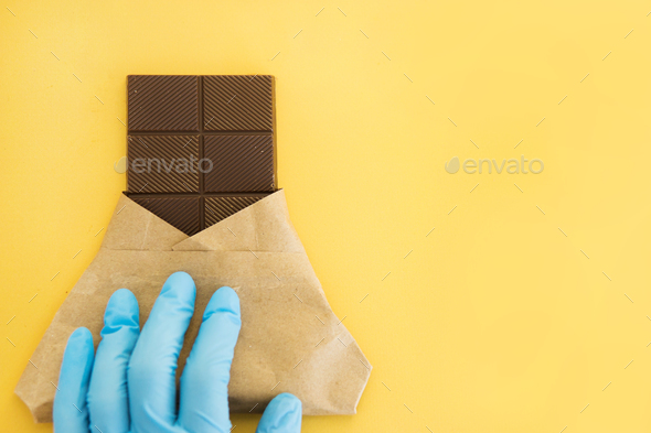 Bar of milk chocolate in a wrapper is held by a hand in a medical glove World chocolate day