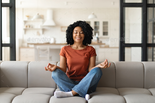 Calm black woman sitting on couch, meditating with closed eyes at home