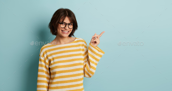 Smiling young Hispanic lady in eyeglasses pointing away in blue studio - Stock Photo - Images