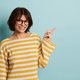 Smiling young Hispanic lady in eyeglasses pointing away in blue studio - PhotoDune Item for Sale