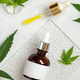 Dropper bottle with blank label near green cannabis leaves on white table. Cosmetic Mockup - PhotoDune Item for Sale