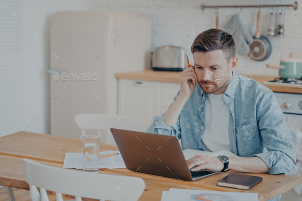 Serious bearded businessman in casual clothes working on laptop while sitting at kitchen table - Stock Photo - Images