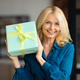 Portrait of happy mature woman sitting on couch at home, holding wrapped gift box, looking and - PhotoDune Item for Sale