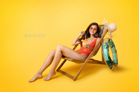Glad millennial mixed race lady in swimsuit sits in deck chair with glass of tropical cocktail - Stock Photo - Images