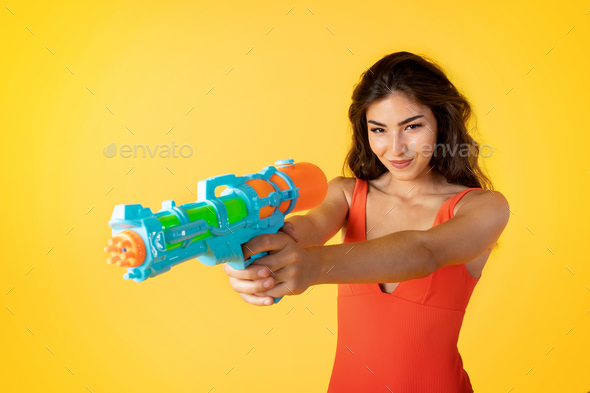 cheerful millennial mixed race lady in swimsuit shoots water pistol has fun, enjoys vacation - Stock Photo - Images